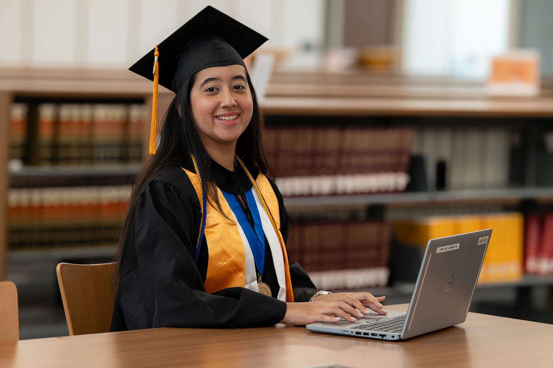 Student wearing graduation cap and gown and using her laptop